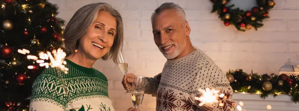 Joyeux couple d'âge moyen en chandails de fête tenant des étincelles et des verres de champagne la veille de Noël, bannière — Photo de stock
