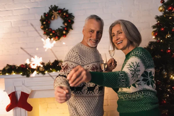 Щаслива зріла пара в святкових светрах дивиться на блискучі блискітки і тримає келихи шампанського напередодні Різдва — стокове фото