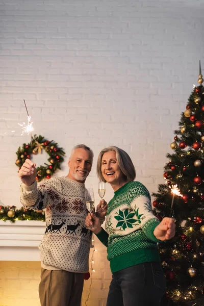 Happy mature couple in sweaters holding glasses of champagne and sparklers on christmas eve — Stock Photo
