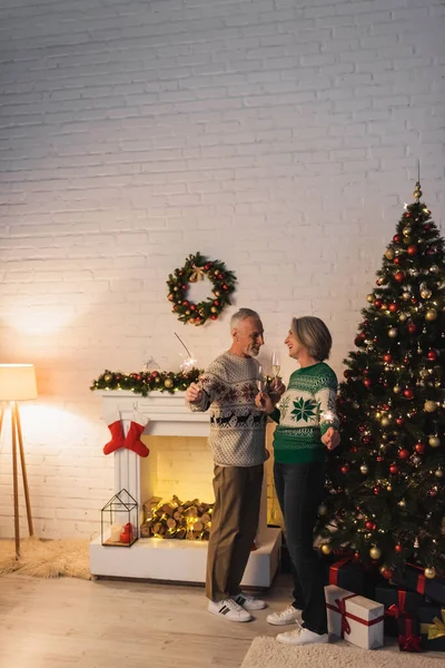 Heureux couple d'âge mûr dans des chandails tenant des verres de champagne et des étincelles le soir de Noël à la maison — Photo de stock