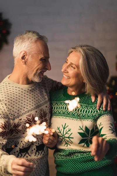 Веселий чоловік середнього віку обіймає дружину, тримаючи іскриста напередодні Різдва — стокове фото
