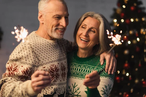 Positiver Mann mittleren Alters umarmt Ehefrau, während er an Heiligabend Wunderkerze hält — Stockfoto