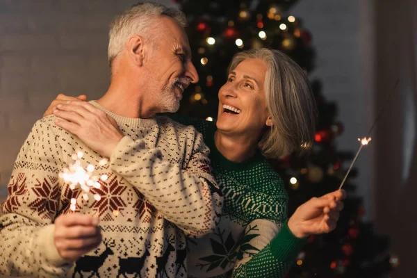 Positive middle aged woman hugging bearded husband while holding bright sparkler on christmas eve — Stock Photo