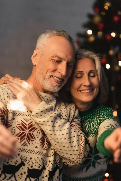 Positivo mulher de meia idade abraçando marido barbudo com olhos fechados enquanto segurando sparkler na véspera de Natal — Fotografia de Stock