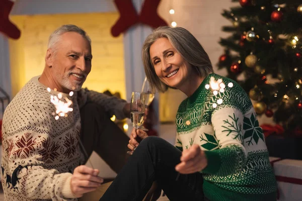 Positives Ehepaar mittleren Alters hält an Heiligabend Sektgläser und glänzende Wunderkerzen in der Hand — Stockfoto