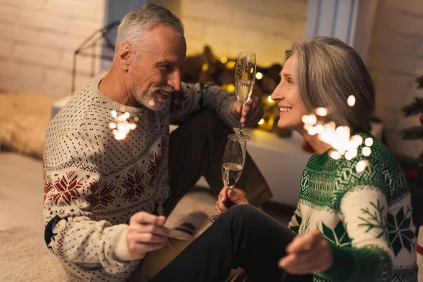Happy middle aged couple holding glasses of champagne and shiny sparklers on christmas eve — Stock Photo