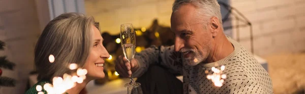 Heureux homme d'âge moyen tenant un verre de champagne et regardant sa femme près des étincelles brillantes la veille de Noël, bannière — Photo de stock