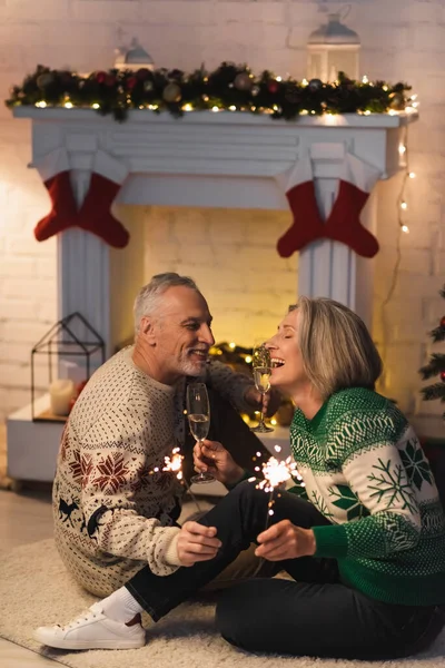 Alegre hombre de mediana edad sosteniendo una copa de champán y mirando a la esposa alegre cerca de brillantes bengalas en la víspera de Navidad - foto de stock