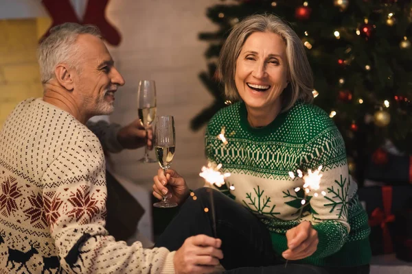 Positive middle aged couple holding glasses of champagne and shiny sparklers on christmas eve — Stock Photo