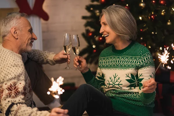 Smiling middle aged couple clinking glasses of champagne and holding shiny sparklers on christmas eve — Stock Photo