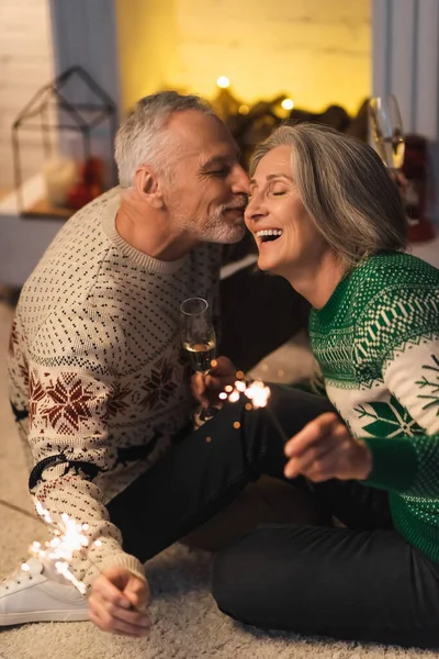 Positive middle aged man kissing wife and holding shiny sparklers on christmas eve — Stock Photo