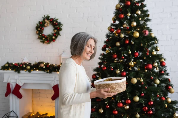 Cheerful and mature woman holding wicker basket near decorated christmas tree at home — Stock Photo