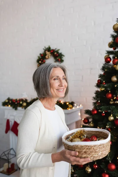Donna felice e matura tenendo cesto di vimini vicino decorato albero di Natale a casa — Foto stock