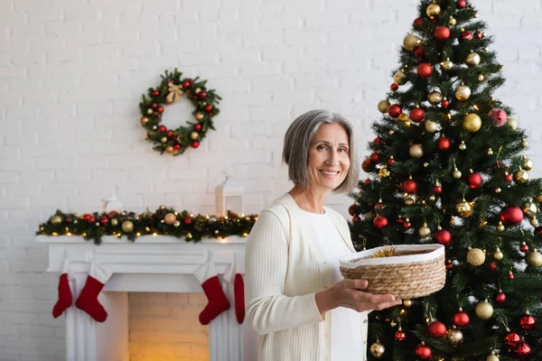 Joyful and mature woman holding wicker basket near decorated christmas tree at home — Stock Photo