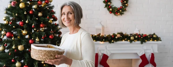 Cheerful and mature woman holding wicker basket near decorated christmas tree at home, banner — Stock Photo