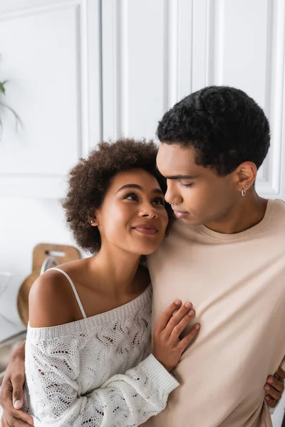Happy african american woman in white openwork sweater looking at boyfriend in kitchen — Stock Photo