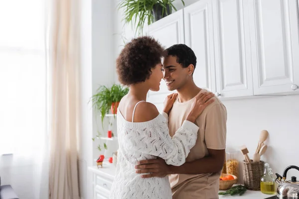 Lockige Afroamerikanerin im weißen durchbrochenen Pullover, die in der Küche neben ihrem glücklichen Freund lächelt — Stockfoto