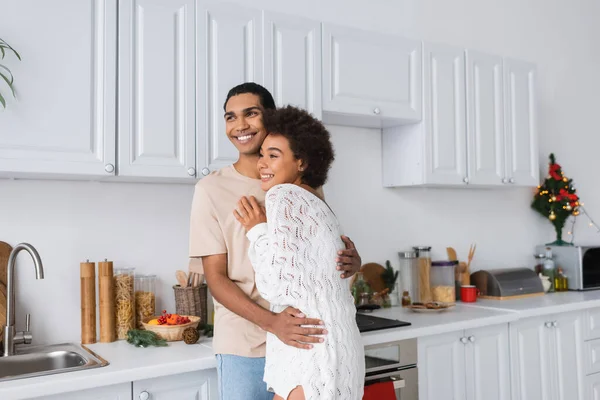 Felice coppia afro-americana che abbraccia e distoglie lo sguardo nella cucina moderna con mobili bianchi — Foto stock