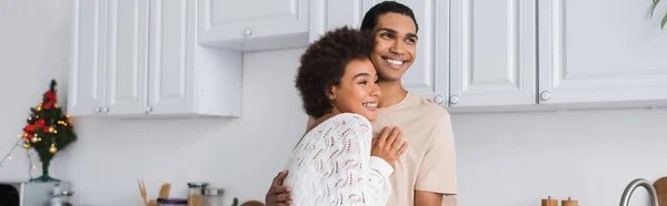 Smiling african american man and woman in white openwork sweater hugging in kitchen, banner — Stock Photo