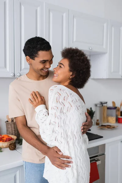 Jeune et heureux couple afro-américain étreignant et se regardant dans la cuisine — Photo de stock