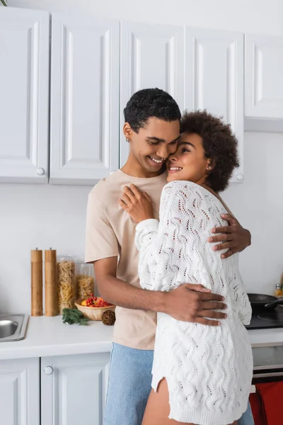 Брюнетка африканская американка в белом трикотажном свитере обнимается с молодым парнем на кухне — стоковое фото
