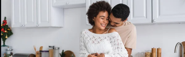Mujer afroamericana feliz con los ojos cerrados riendo cerca de novio en la cocina, pancarta - foto de stock