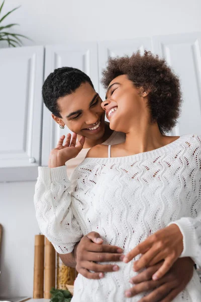 Sorridente donna afroamericana in maglione bianco traforato che tocca il viso dell'uomo abbracciandola in cucina — Foto stock
