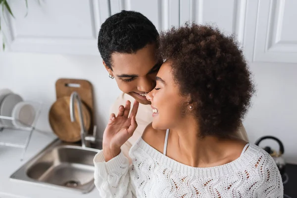 Sensual african american woman in white knitted sweater touching face of smiling boyfriend in kitchen — Stock Photo