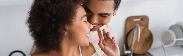 Curly african american woman touching face of smiling boyfriend in kitchen, banner — Stock Photo