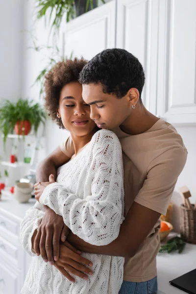 Jovem homem americano africano com piercing abraçando mulher sorridente em suéter branco openwork na cozinha — Fotografia de Stock
