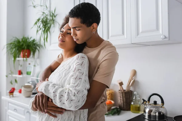 Morena africano americano hombre abrazando novia en blanco suéter mirando hacia otro lado en cocina - foto de stock