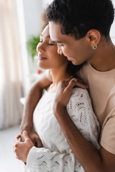 Blurred african american woman in white sweater smiling with closed eyes near boyfriend in kitchen — Stock Photo