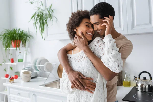 Alegre africana americana mujer en blanco punto suéter abrazando con joven novio en cocina - foto de stock