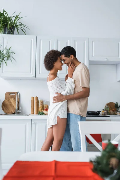 Vista lateral de mujer afroamericana en suéter blanco abrazando con novio en cocina - foto de stock