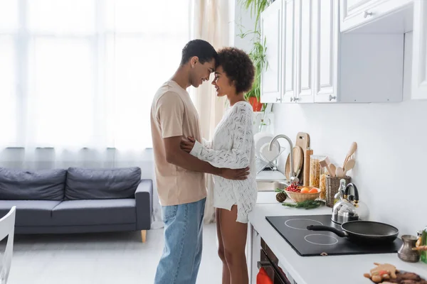 Vista lateral de hombre afroamericano feliz en jeans y mujer en cocina moderna - foto de stock