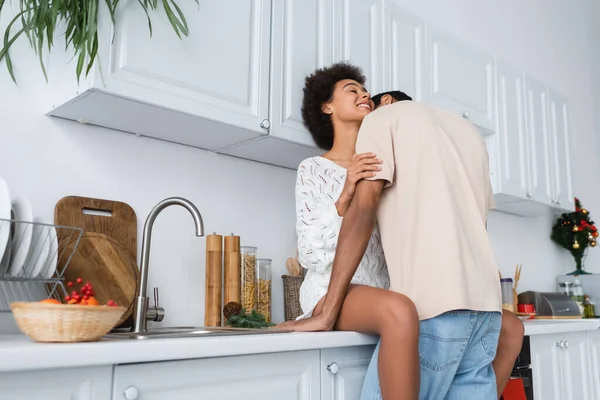 African american man hugging happy girlfriend in white sweater sitting on worktop in kitchen — Stock Photo