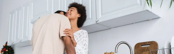 Low angle view of young african american man hugging cheerful girlfriend in kitchen, banner — Stock Photo