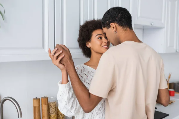 Pleased african american woman in white sweater holding hands with boyfriend in kitchen — Stock Photo