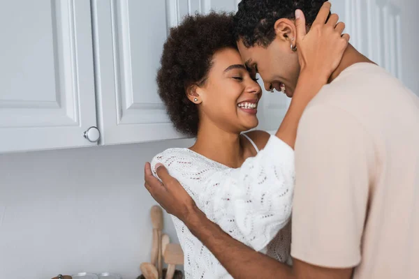 Cheerful african american woman with closed eyes hugging boyfriend near white furniture in kitchen — Stock Photo