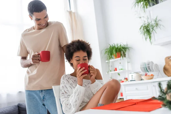 Joven afroamericano hombre sonriendo cerca sexy novia beber café de la mañana con los ojos cerrados - foto de stock