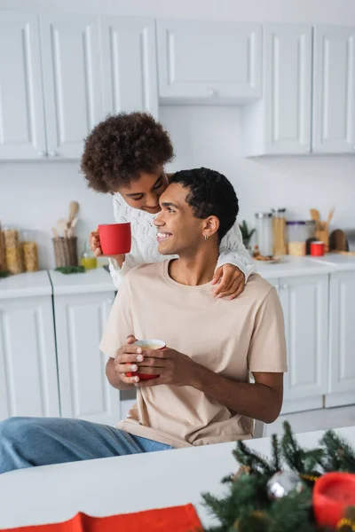 Curly african american woman with red cup near smiling boyfriend in kitchen — Stock Photo