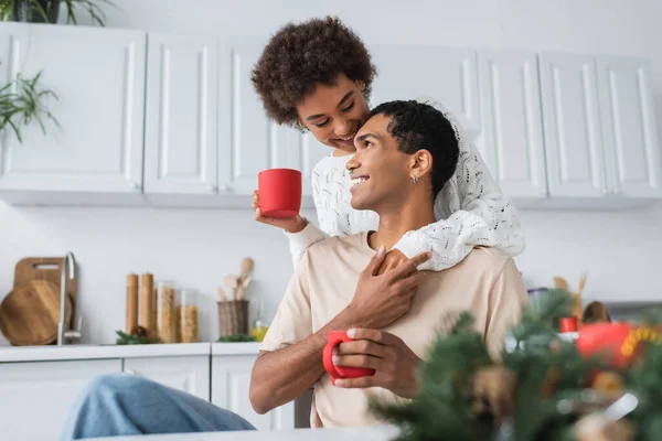 Afrikanisch-amerikanische Frau und ihr lächelnder Freund mit roten Tassen Händchen haltend und lächelnd in der Küche — Stockfoto