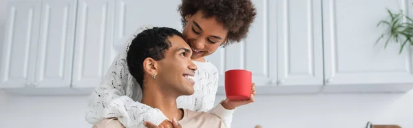 Joyful african american woman in white sweater holding red cup near happy man in ktchen, banner — Stock Photo