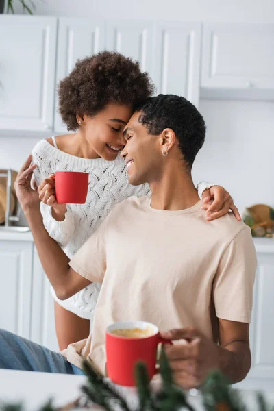 Joven y feliz pareja afroamericana sosteniendo tazas rojas y abrazándose en la cocina - foto de stock