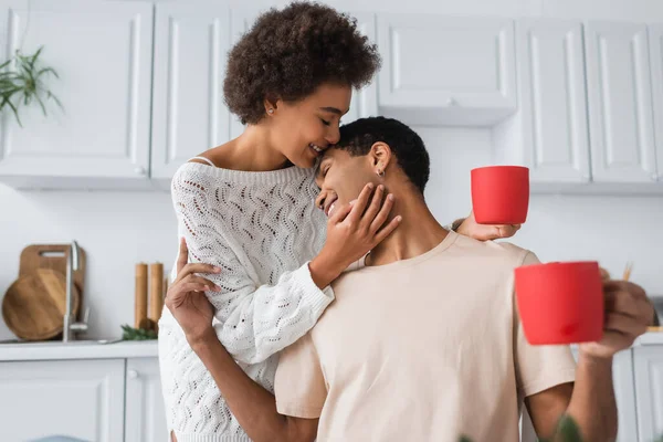 Heureux couple afro-américain avec des tasses rouges étreignant dans la cuisine — Photo de stock