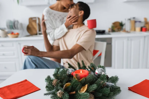Couronne de Noël avec bougie près de serviettes rouges et couple afro-américain embrassant sur fond flou — Photo de stock
