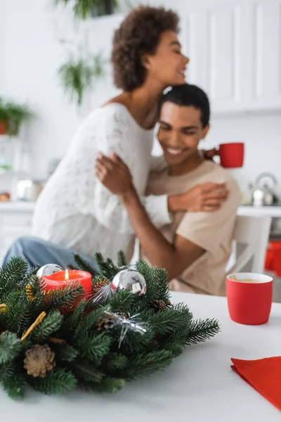 Couronne de Noël décorée de bougies et de boules près du couple afro-américain flou embrassant dans la cuisine — Photo de stock