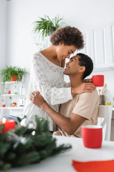 Smiling african american woman with red cup hugging young brunette boyfriend in kitchen — Stock Photo