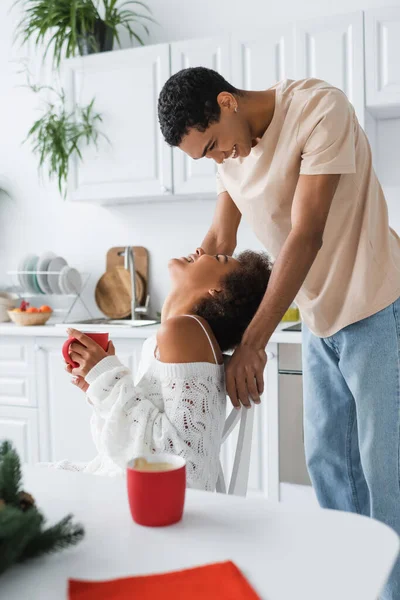 Mujer afroamericana emocionada en suéter de punto blanco sosteniendo taza roja y mirando novio feliz - foto de stock
