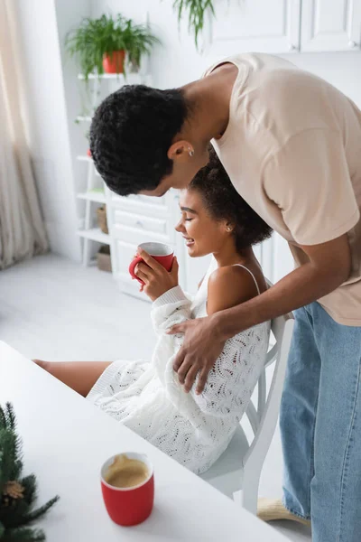 Young african american man near smiling girlfriend sitting with cup of cocoa in kitchen — Stock Photo
